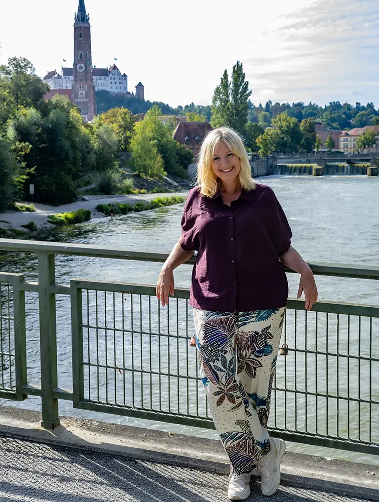 Eine Frau posiert lächelnd am Flussufer in Landshut, mit der Burg im Hintergrund, als mögliche Stadtratskandidatin für die Kommunalwahl 2026.